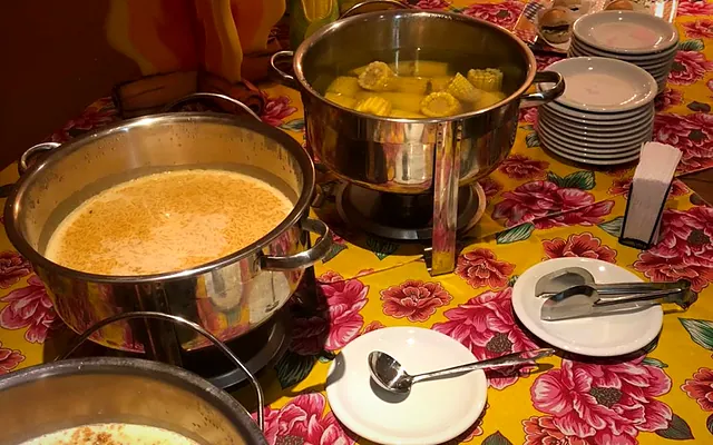 Mesa do buffet delivery com rechauds, milho cozido e doces servidos sobre toalha de chita